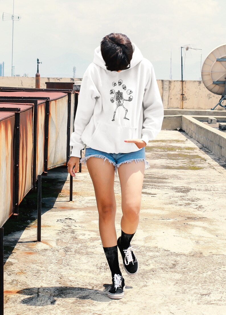 May include: A white hoodie featuring a graphic of a skeleton juggling skulls. The person wearing the hoodie is walking on a rooftop.