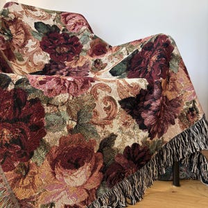 May include: A floral tapestry throw blanket draped over a chair. The blanket features a pattern of large burgundy and pink roses with green leaves on a beige background. The edges are finished with a black and white fringe.