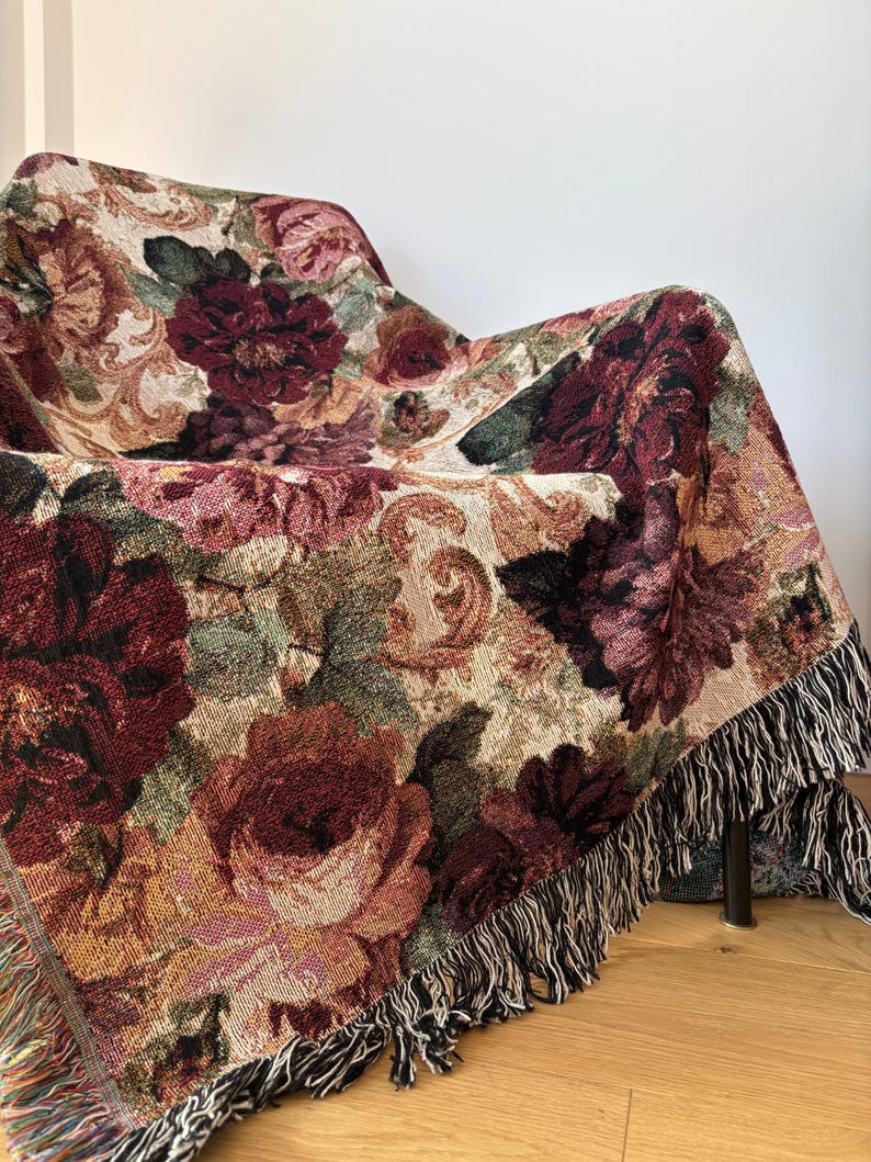 May include: A floral tapestry throw blanket draped over a chair. The blanket features a pattern of large burgundy and pink roses with green leaves on a beige background. The edges are finished with a black and white fringe.