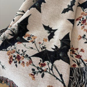 May include: Beige throw blanket with a repeating pattern of black bats and floral accents in shades of orange, red, and green. The blanket has a fringed edge.