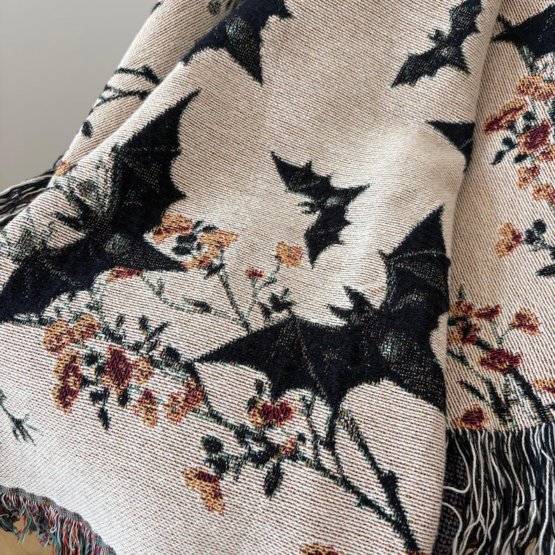 May include: Beige throw blanket with a repeating pattern of black bats and floral accents in shades of orange, red, and green. The blanket has a fringed edge.