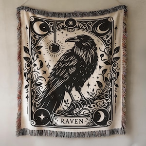 May include: A woven throw blanket featuring a black raven illustration. The design includes moons, stars, and botanical elements, with the word "RAVEN" at the bottom. The blanket has a cream background and fringed edges.