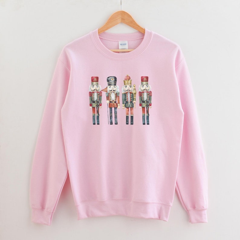 May include: A light pink sweatshirt featuring a watercolor illustration of four toy nutcracker soldiers. The nutcrackers are dressed in red, black, green, and gold uniforms. The sweatshirt is hanging on a wooden hanger.