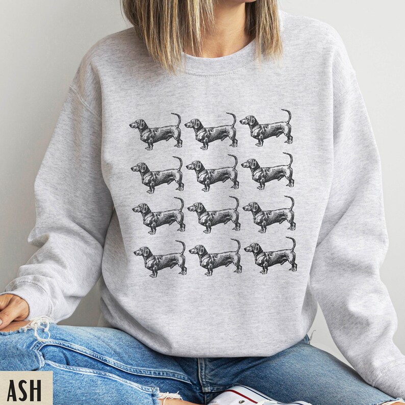 Peut inclure: Un sweat-shirt gris avec un motif r&eacute;p&eacute;t&eacute; de chiens teckels noirs. Les chiens sont tous tourn&eacute;s dans la m&ecirc;me direction et sont dans diff&eacute;rentes poses.