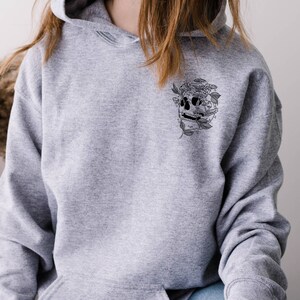 May include: A gray hoodie featuring a black and white skull design with mushrooms and leaves. The graphic is on the upper left chest area. The hoodie has a drawstring hood and a kangaroo pocket.