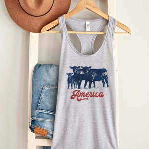 May include: Gray tank top with a graphic of four black cows and the text "America" in red, white, and blue. The tank top is hanging on a wooden hanger.
