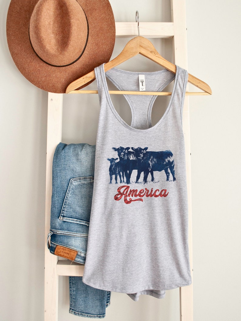 May include: Gray tank top with a graphic of four black cows and the text "America" in red, white, and blue. The tank top is hanging on a wooden hanger.