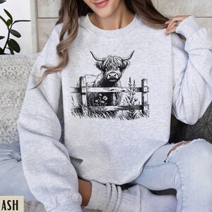 Puede incluir: Una sudadera gris con una ilustraci&oacute;n en blanco y negro de una vaca de las tierras altas mirando por encima de una valla. La vaca est&aacute; de pie en un campo con flores.