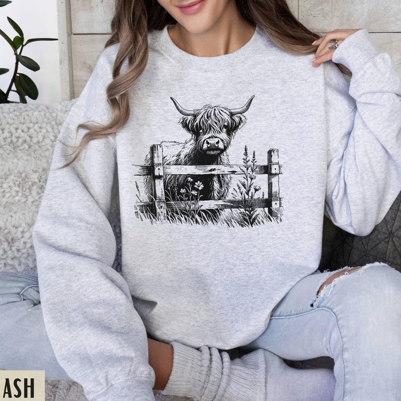 Puede incluir: Una sudadera gris con una ilustraci&oacute;n en blanco y negro de una vaca de las tierras altas mirando por encima de una valla. La vaca est&aacute; de pie en un campo con flores.