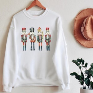 May include: White crewneck sweatshirt featuring a watercolor illustration of four toy nutcracker soldiers. The nutcrackers are in red, black, and green uniforms with gold accents. The sweatshirt is hanging on a wooden hanger.