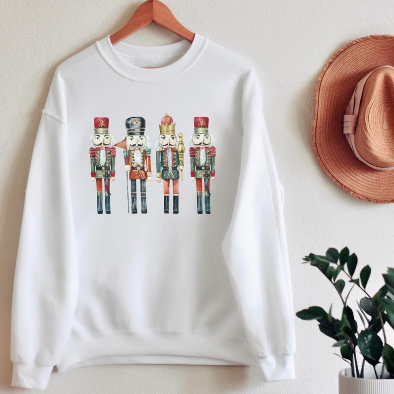 May include: White crewneck sweatshirt featuring a watercolor illustration of four toy nutcracker soldiers. The nutcrackers are in red, black, and green uniforms with gold accents. The sweatshirt is hanging on a wooden hanger.