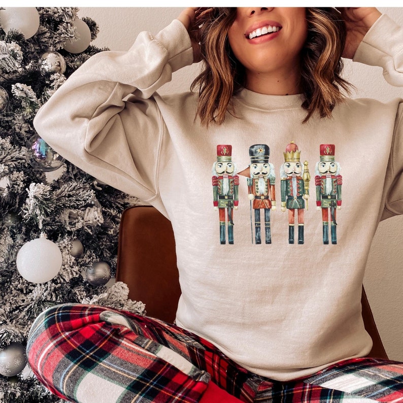 May include: Beige sweatshirt featuring a watercolor illustration of four toy nutcrackers. The nutcrackers are in various colors, including red, green, and gold, and are standing in a row. The sweatshirt is a neutral color, and the overall theme is holiday-themed.