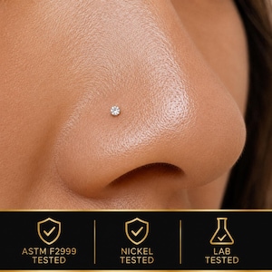 May include: A small, round, clear gemstone nose stud set in a metallic base. Below the nose, a black banner displays three icons with checkmarks and the text: ASTM F2999 TESTED, NICKEL TESTED, and LAB TESTED, indicating the jewelry's safety standards.