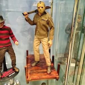 May include: A figure of Jason Voorhees wearing a hockey mask, khaki pants, and a brown button-up shirt, holding a machete. The figure stands on a wooden dock. Other figures are visible in a glass display case.
