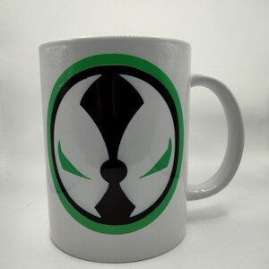 May include: White ceramic mug featuring a green and black graphic design. The design is a stylized, symmetrical emblem with a circular border. The mug has a curved handle and is suitable for hot or cold beverages.