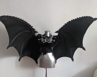 May include: A black costume with large, pointed wings, displayed on a silver mannequin. The wings have a textured, jagged edge. The bodice is embellished with silver beads and black feathers, creating a dramatic, theatrical look.
