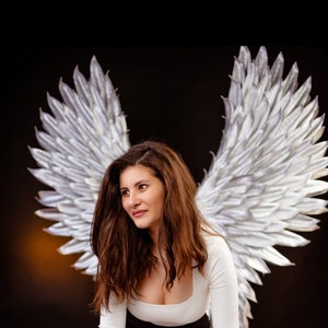 Op de afbeelding: Een vrouw met lang bruin haar, die een wit overhemd met lange mouwen en een zwarte broek draagt, poseert op een witte kubus. Ze draagt zilveren engelenvleugels.
