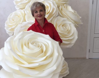 May include: A woman in a red shirt stands in front of a wall decorated with large, white paper roses. The roses are arranged in a cluster, with the largest rose in the center.