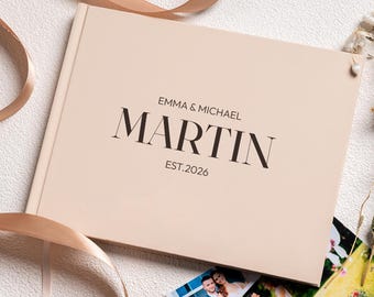 May include: A beige rectangular photo album with a hard cover featuring the printed text EMMA & MICHAEL MARTIN EST.2026 in dark brown lettering. The album is accented by a light brown satin ribbon and a small cluster of dried white flowers.
