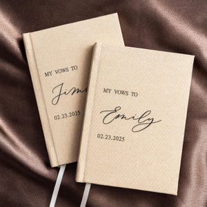 May include: Two beige linen-covered vow books with the text "MY VOWS TO" and names in elegant script. The date "02.23.2025" is printed below the names. White ribbon bookmarks are visible.