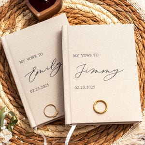 Peut inclure: Deux carnets de vœux en lin beige avec du texte noir. L'un porte l'inscription MY VOWS TO Emily 02.23.2025, l'autre MY VOWS TO Jimmy 02.23.2025. Chaque carnet possède un marque-page en ruban blanc et une bague en métal doré sur la couverture.
