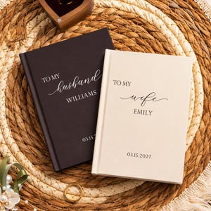 May include: A dark brown journal with white text reading TO MY husband WILLIAMS 03.15.2027 and a cream journal with black text reading TO MY wife EMILY 03.15.2027. Both journals feature a hard cover and are positioned side by side.
