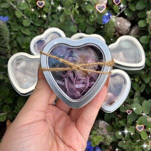 May include: Five heart-shaped metal tins with clear lids. Each tin contains a different crystal or stone wrapped in purple and white tissue paper. The tins are arranged in a semi-circle with the largest tin in the center.