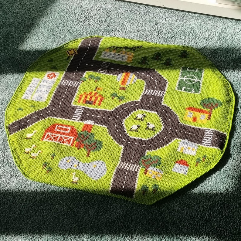 May include: A green crocheted playmat with a town scene. The mat features a road with a roundabout, houses, a tent, a hot air balloon, and sheep.