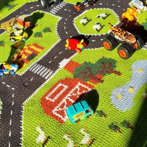 May include: A green and gray play mat with a road and parking lot design.  There are toy cars and trucks on the mat, including a red airplane, a blue van, a green monster truck, and a yellow monster truck.