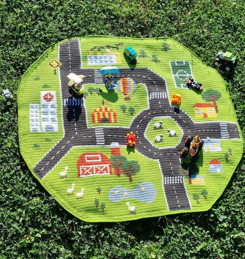 May include: A green crocheted play mat with a road and city design. The mat features a variety of small toy cars and trucks, including a red car, a blue truck, and a green truck. The mat is perfect for children who love to play with cars and trucks.