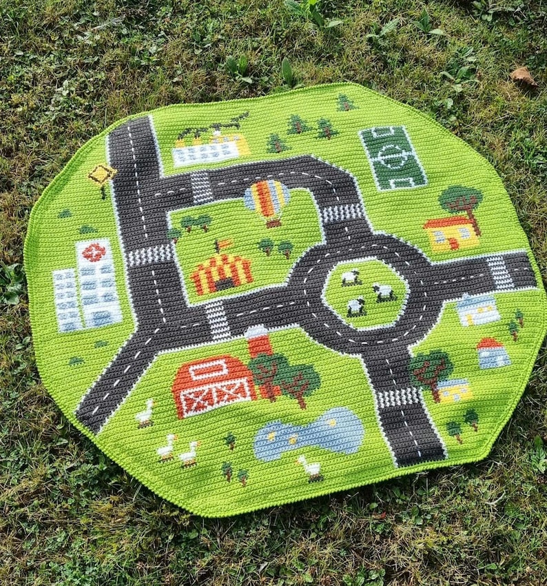 May include: A green crocheted playmat with a road and city design. The playmat features a roundabout, a soccer field, a farm, a circus tent, and a hot air balloon.