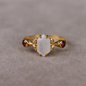 Puede incluir: Un anillo de oro con una piedra lunar hexagonal central flanqueada por dos pequeñas piedras granates rojas redondas. El engaste incluye delicados detalles metálicos en forma de hoja que rodean la piedra central sobre un aro fino.