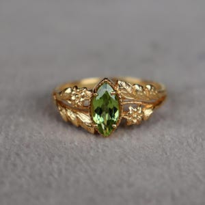 Könnte beinhalten: Ein goldener Ring mit einem zentralen, marquise-geschliffenen grünen Edelstein. Die Ringschiene ist mit filigranen Metallarbeiten verziert, die kleine Blumen und Blätter auf beiden Seiten des Steins darstellen und ein detailliertes florales Motiv bilden.