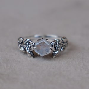 May include: A silver ring featuring a central hexagonal moonstone set in prongs. The band is crafted with detailed metalwork depicting small flowers, crescent moons, and leaves extending from the center stone along the sides of the ring.