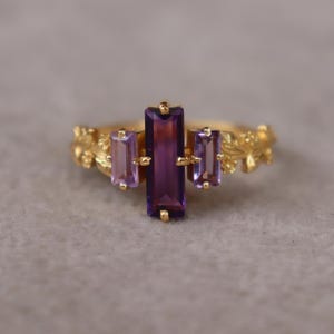 May include: A gold ring featuring a central vertical baguette-cut purple amethyst flanked by two smaller vertical baguette-cut light purple amethysts. The gold band is detailed with a raised relief pattern of small leaves and flowers.