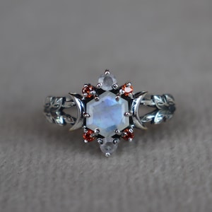 May include: A silver ring featuring a central hexagonal moonstone, flanked by two crescent moon shapes and two small round clear stones. Four vibrant orange-red round stones are set around the center. The band has a detailed leaf motif design.