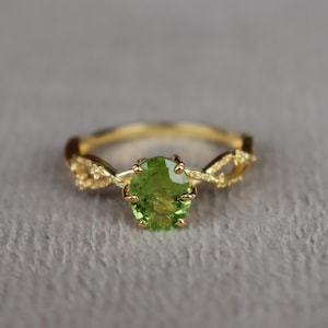 Könnte beinhalten: Ein Goldring mit einem oval geschliffenen, leuchtend lindgrünen Edelstein in einer Krappenfassung. Die Ringschiene ist mit einem strukturierten, rankenartigen Design gestaltet, das sich auf beiden Seiten des Mittelsteins windet und verzweigt.