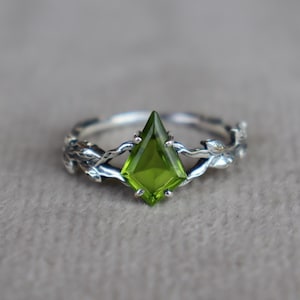 May include: A silver ring featuring a kite-shaped, vibrant green gemstone set in the centre. The band is crafted to resemble intertwined branches with small leaves, providing a textured, organic appearance that frames the central stone securely.