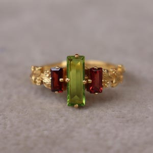 May include: A gold ring featuring a central vertical baguette-cut lime green gemstone flanked by two smaller vertical baguette-cut deep red gemstones. The ring band is crafted with a detailed relief pattern of leaves and small floral buds.