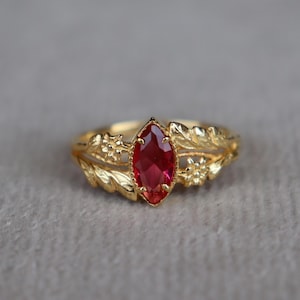 May include: A gold ring featuring a central marquise-cut red gemstone. The band is designed with intricate leaf and small flower motifs on both sides of the stone, creating a detailed botanical appearance around the vibrant red centre piece.