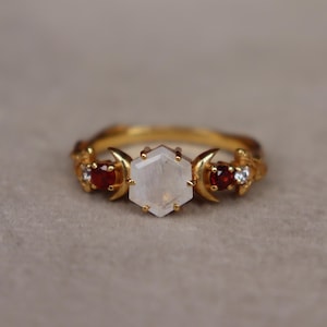 Op de afbeelding: Een gouden ring met een centrale zeshoekige maansteen geflankeerd door twee halvemaanmotieven. Elke kant bevat een ronde rode granaat en een kleine heldere steen, gezet in een ringband met bladvormige details aan de buitenranden.
