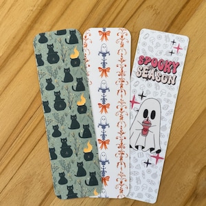 May include: Three rectangular bookmarks with distinct designs. The first features black cats in cauldrons on a sage green background. The second has a vertical pattern of ghosts and orange bows. The third displays the text SPOOKY SEASON with a ghost.