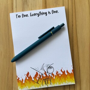 May include: A white notepad with the text "I'm fine. Everything is fine." and a cartoon of a person in a burning room. A teal pen rests on the notepad. The notepad has a yellow and orange flame design at the bottom.