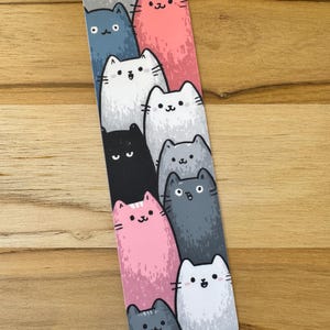 May include: A rectangular bookmark featuring a repeating pattern of cartoon cats in various colors, including pink, gray, white, black, and blue. The cats have simple, smiling faces and are arranged vertically.