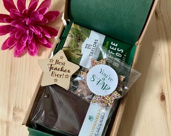 May include: A green gift box with a brown lid is open and filled with a variety of treats. The box contains a chocolate bar, a bag of coffee, a star-shaped chocolate treat with sprinkles, and a wooden star with the text "Best Teacher Ever!" on it. The box is sitting on a wooden surface.