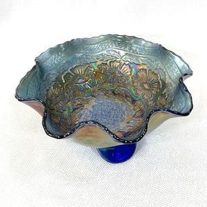 May include: An iridescent blue glass bowl with a ruffled edge and a blue pedestal base. The bowl features a floral pattern in shades of gold and green. The bowl is likely vintage or antique.