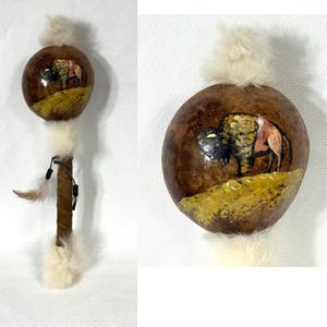 May include: A handcrafted rattle with a round, brown gourd head featuring a painted bison scene. The handle is wrapped in brown leather and adorned with white feathers and a small feather accent. The rattle is a traditional Native American instrument.