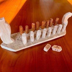 Puede incluir: Un juego de barco vikingo de madera con piezas de madera tallada. El barco es de color claro con un diseño detallado, y las piezas del juego son cilíndricas. También se incluyen dos pequeñas baldosas de madera con símbolos grabados.