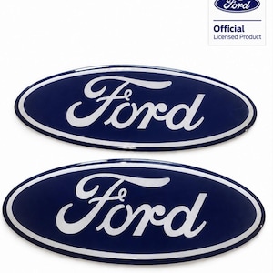 Op de afbeelding: Twee ovale Ford-emblemen met een donkerblauwe achtergrond, witte letters en een witte rand. Een klein vierkant label in de rechterbovenhoek toont het Ford-logo en de tekst Official Licensed Product in het zwart en blauw.