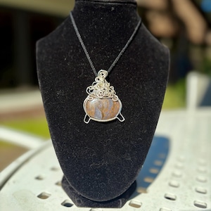 May include: A handcrafted pendant necklace featuring a wire-wrapped stone in the shape of a cauldron. The stone has brown and tan hues. The pendant hangs from a black cord necklace, displayed on a black velvet bust.
